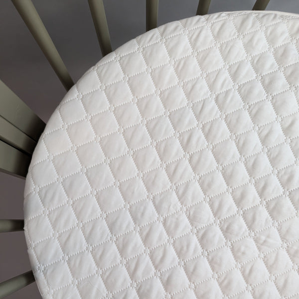 A close-up image of a white, quilted mattress protector with a diamond pattern on a Stokke Sleepi Mini model bassinet.
